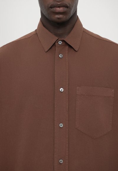 Chemise marron à boutons, fabriquée en tissu lisse, avec un col, une fermeture à six boutons et une poche poitrine gauche.