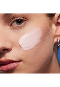 Fuktighetskräm applicerad på en kind, slät, glänsande, ljusrosa i färgen, med en krämig konsistens. Huden verkar frisk och välvårdad.