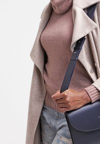 Persona vestida con un abrigo beige y un suéter de cuello alto malva, sosteniendo un bolso de cuero azul oscuro con una correa gruesa.