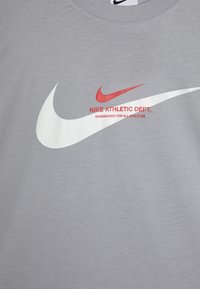 Pilka marškinėliai su ryškiu baltu "Nike" logotipu, mažesniu raudonu logotipu ir juodu tekstu "NIKE ATHLETIC DEPT." ant krūtinės.