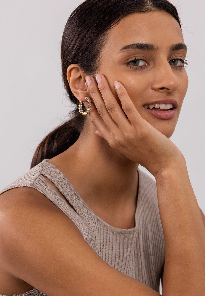 Goldfarbene Creolen mit gedrehter Optik. Das Model trägt ein beiges, geripptes Oberteil und posiert mit der Hand im Gesicht, um die Ohrringe zu zeigen.