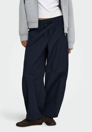 Personne portant un pantalon large bleu marine foncé avec une taille à cordon, des chaussures marron, un haut blanc et une veste grise zippée.