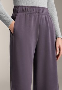 Fialové kalhoty s elastickým pasem a hladkým povrchem. Boční kapsy a uvolněný střih. Částečně viditelný šedý top s dlouhým rukávem s vroubkovaným vzorem.