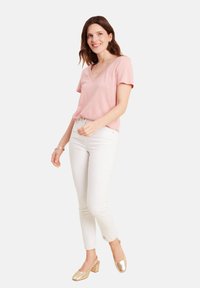 Hellrosa V-Ausschnitt-T-Shirt aus weichem Stoff, kombiniert mit weißen Slim-Fit-Jeans. Getragen mit goldenen Blockabsatzschuhen und runden Ohrringen.
