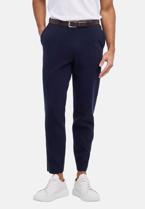 Mann, der marineblaue Slim-Fit-Hosen mit braunem Gürtel, weißem T-Shirt und weißen Sneakers trägt, steht mit teilweise in den Taschen steckenden Händen.