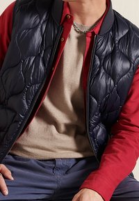 Gilet trapuntato blu su una maglietta rossa a maniche lunghe, abbinato a un maglione beige colletto rotondo e pantaloni blu. Tessuto liscio con un design a strati.