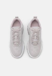 Et par beige Guess sneakers med præget logo mønstre, snørebånd foran og hvide tykke såler på en ensfarvet hvid baggrund.