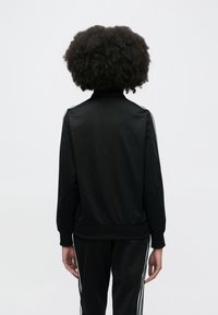 Must spordijakk kõrge kraega, siledast materjalist ja soonikservadega. Varrastel valged triibud ning sobivad püksid samas stiilis.