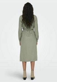 Abito a maniche lunghe di colore verde oliva con vita segnate da una cintura, realizzato in tessuto strutturato. Presenta polsini con bottoni e arriva sotto il ginocchio. Stivali chunky beige.