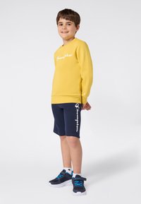 Un garçon souriant portant un sweatshirt jaune Champion, un short bleu marine avec un logo blanc, des chaussettes blanches et des baskets bleu marine avec des lacets bleus, se tenant devant un fond blanc.