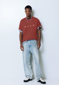 T-shirt oversize couleur rouille avec des manches rayées, comportant un texte brodé blanc. Jeans bleu clair à jambes larges et chaussures noir et blanc.