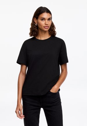 Femme aux cheveux bouclés portant des boucles d'oreilles argentées en forme de créoles, un t-shirt noir uni à manches courtes et un pantalon noir, debout avec une main dans sa poche.