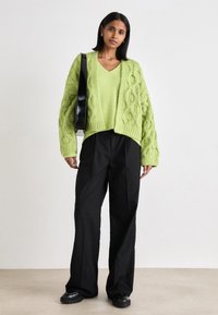 Λάιμ πράσινο πλεκτό oversize πουλόβερ με σχέδιο καμπάνας, συ�νδυασμένο με μαύρα παντελόνια με φαρδιά πόδια και μαύρα χοντρά παπούτσια, κρατώντας μια μαύρη τσάντα.