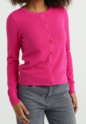 Woman wearing a fuchsia textured button-up cardigan with long sleeves and gray jeans, with hand resting near pocket.