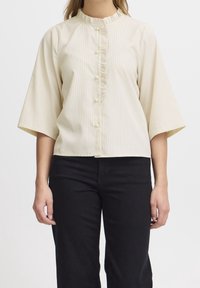 Krämfärgad skjorta med vertikala ränder, halvlånga utsvängda ärmar, en rynkad knapplist och små knappar. Matchad med svarta byxor.