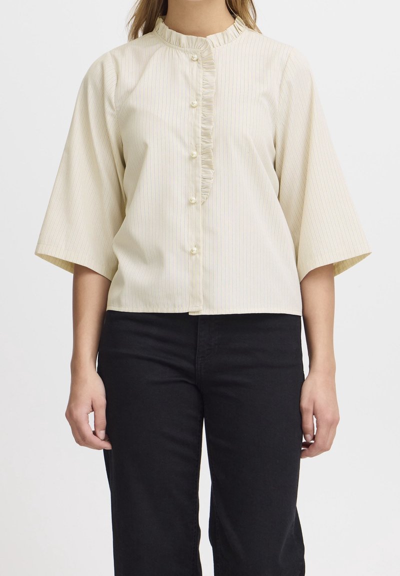 Krämfärgad skjorta med vertikala ränder, halvlånga utsvängda ärmar, en rynkad knapplist och små knappar. Matchad med svarta byxor.