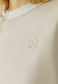 Beige top met een textuurstof kraag en een gegraveerd "OUI" logo op de voorkant, met een glad, minimalistisch ontwerp.