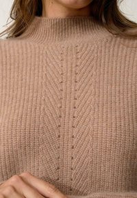 Beige gebreide trui met een hoge hals, voorzien van een ribbelstructuur en verticale lijnen, die een zachte en comfortabele uitstraling benadrukken.