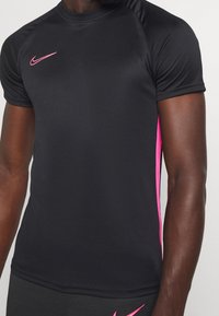 Sort trænings T-shirt med korte ærmer, der har pink accenter og et lille logo nær halsudskæringen. Glat, tætsiddende design.