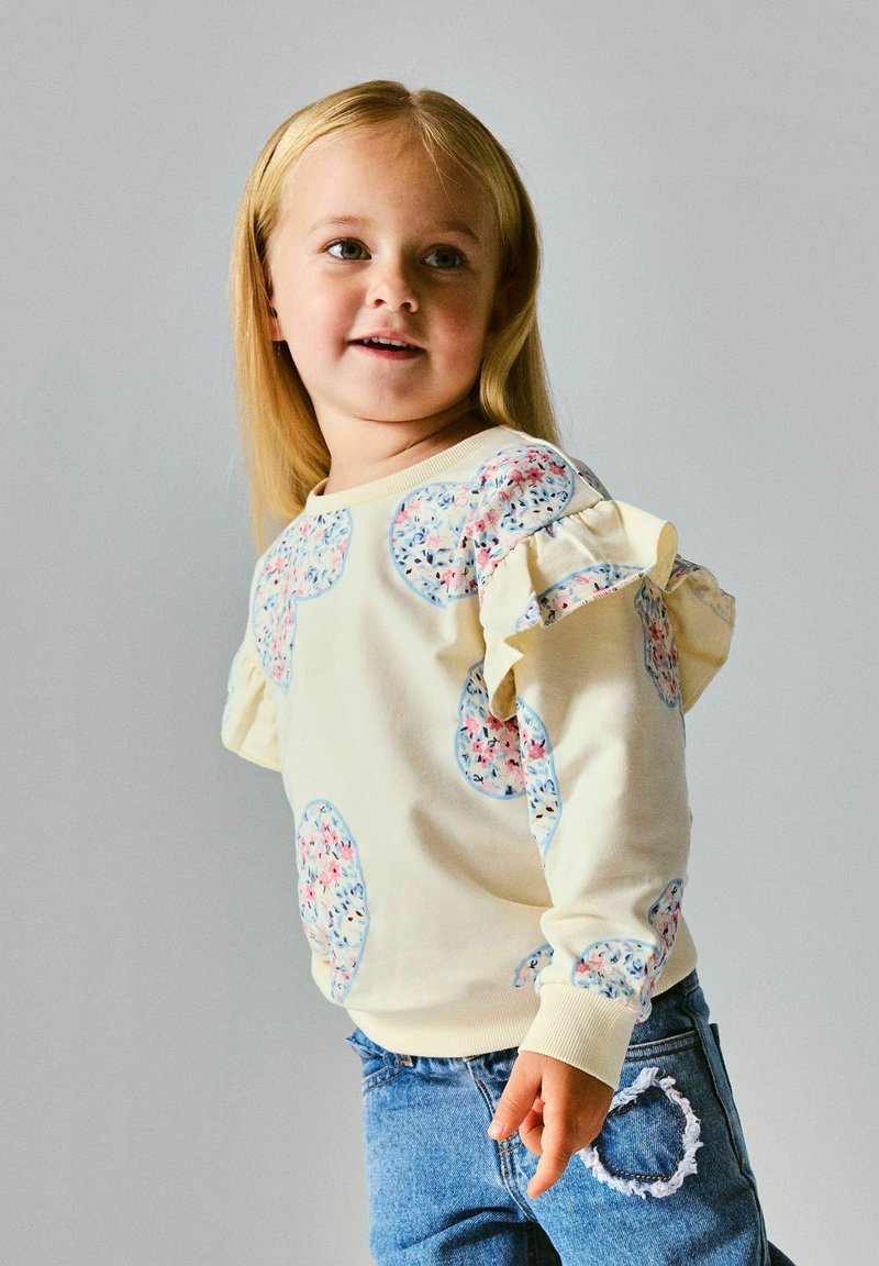 Enfant blond souriant portant un pull crème avec des pois floraux et des manches à volant, associé à un jean bleu avec un patch en forme de cœur.