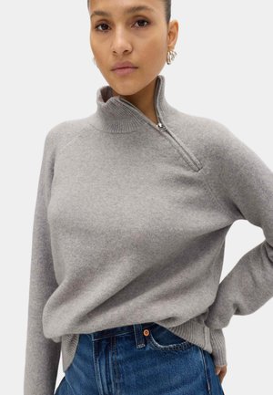 Mujer vistiendo un suéter gris claro con cremallera y cuello alto, y jeans de mezclilla azul, posando con una mano en la cadera frente a un fondo liso.