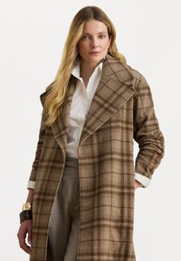 Cappotto di lana a quadri marrone con ampie revers, maniche lunghe e una vestibilità comoda. Indossato sopra una camicia bianca con bottoni e pantaloni grigi.