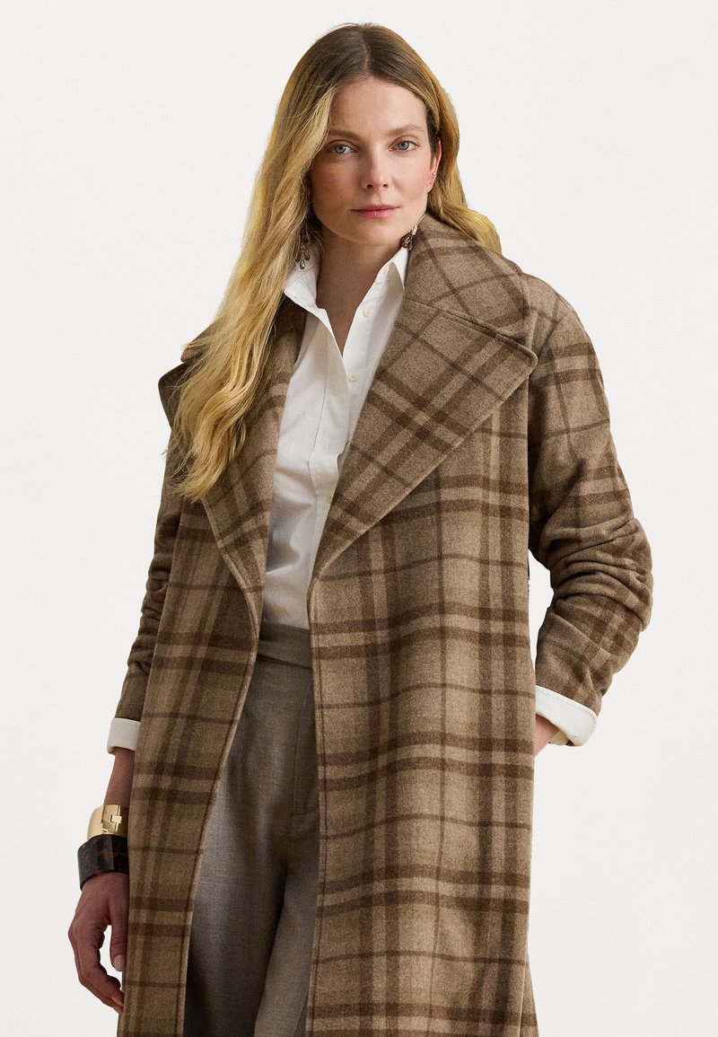 Cappotto di lana a quadri marrone con ampie revers, maniche lunghe e una vestibilità comoda. Indossato sopra una camicia bianca con bottoni e pantaloni grigi.