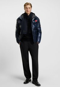 Giacca puffer blu marino con finitura lucida, toppa rossa con logo, abbinata a una sciarpa nera, un maglione nero, pantaloni su misura e scarpe nere.
