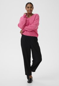 Suéter rosa de punto borroso con cuello en V y dobladillo acanalado, combinado con pantalones negros de corte amplio y zapatos negros. Fondo limpio.