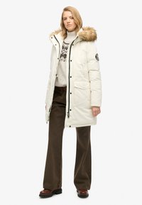 Hvid pufferjakke med en faux fur hætte, lynlås og store frontlommer. Båret over en mønstret sweater og brune vide bukser.