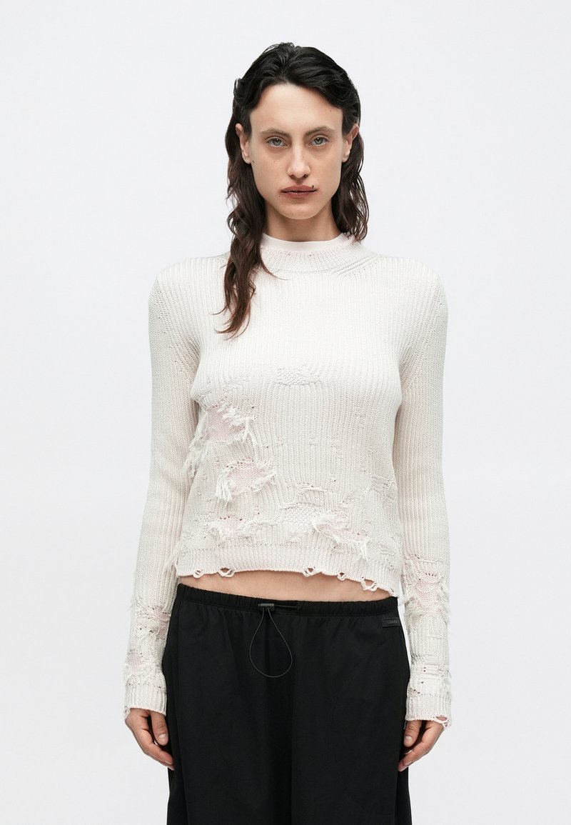 Pulover crem tricotat cu guler înalt, cu detalii uzate și margini destrămate, asortat cu pantaloni negri cu picior larg.