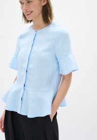 Svetle modrá blůza s krátkým rukávem a kulatým výstřihem, s knoflíky vpředu a peplum spodní částí. Vyrobena z texturované, lehké tkaniny.
