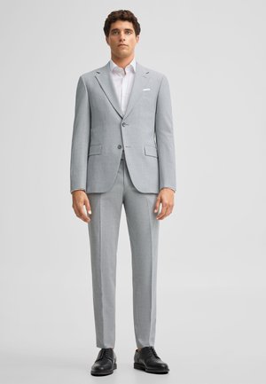 Mężczyzna stoi ubrany w jasnoszary garnitur z białą koszulą i czarnymi eleganckimi butami na jednolitym jasnym tle.