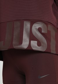 Maroonowa sportowa bluzka z rypsowaną fakturą i dużym nadrukiem "JUST" w błyszczącym materiale; zestawiona z maroonowymi legginsami z małym szarym logo Nike.