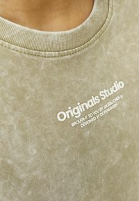 Olive žalia sportinė palaidinė su gilaus kaklo iškilimais ir lygia tekstūra, ant kurios yra baltai spausdintas tekstas „Originals Studio“ ir papildomi detalės.