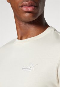 Tricou din bumbac de culoare crem cu guler rotund, având un logo Puma brodat alb pe piept și o textură netedă.