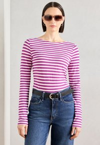 Gestreiftes langärmliges Hemd in Lila und Weiß, kombiniert mit blauen Jeans und einem schwarzen Gürtel. Das Modell trägt dunkle Sonnenbrillen und silberne Creolen.