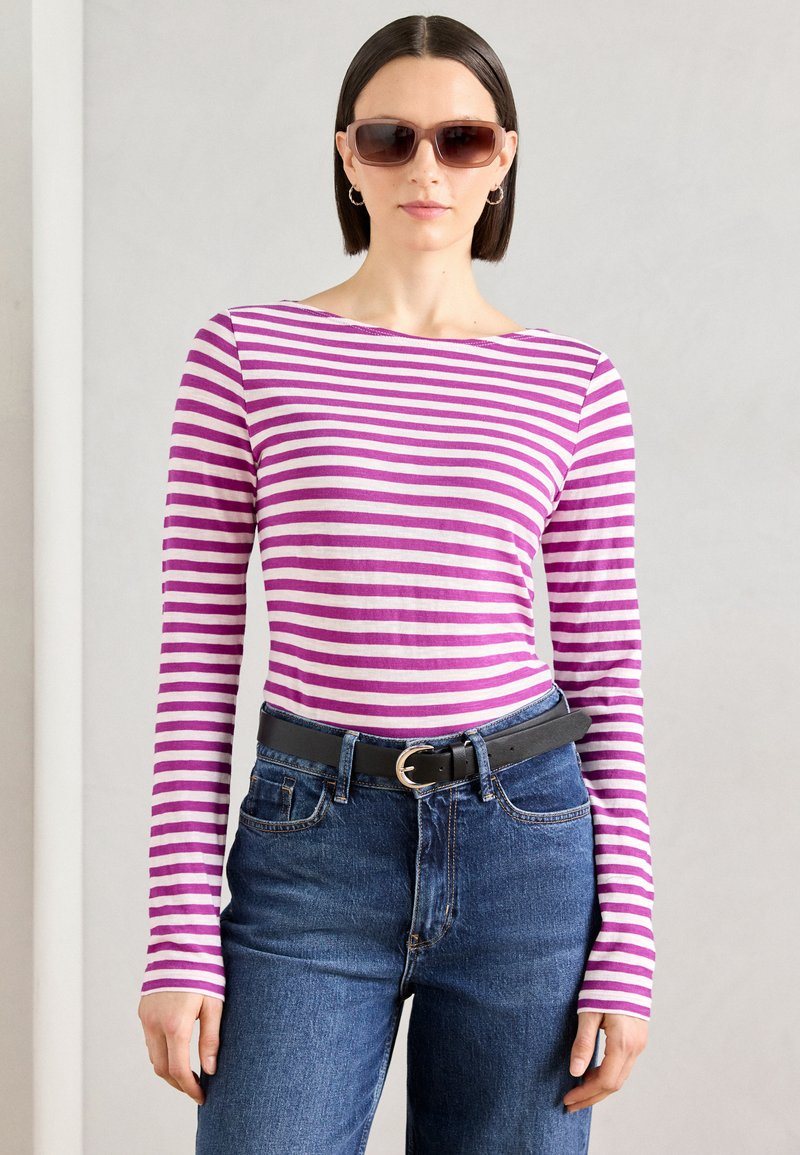 Gestreiftes langärmliges Hemd in Lila und Weiß, kombiniert mit blauen Jeans und einem schwarzen Gürtel. Das Modell trägt dunkle Sonnenbrillen und silberne Creolen.