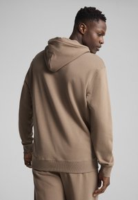Sudadera marrón con capucha, fabricada en un tejido suave, de corte relajado y con puños de canalé. Presenta un diseño liso con toques mínimos, ideal para el uso casual.