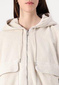 Närbild av en person som bär en krämfärgad kordflodzip-up hoodie med huva och framfickor, som visar bara läpparna och den nedre delen av ansiktet.