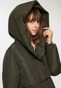 Giacca imbottita verde oliva con un grande cappuccio, dotata di chiusura con zip e polsini a costine. Il materiale appare lucido e texturizzato.