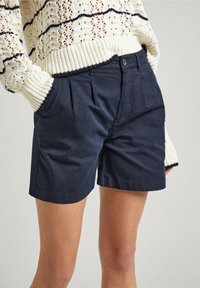 Pantaloni corti in cotone blu navy con pieghe e tasche laterali, tagliati sopra il ginocchio. Abbinati a un maglione in maglia color crema con un motivo testurizzato.