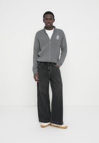 Γκρι ριγωτό πουλόβερ με φερμουάρ και λεπτομέρεια λογότυπου, συνδυασμένο με μαύρο τζιν φαρδιάς γραμμής και μπεζ αθλητικά παπούτσια, σε λευκό φόντο.