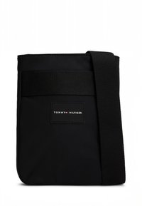 Schwarze Umhängetasche mit breitem, verstellbarem Schulterriemen und "Tommy Hilfiger" Logo-Patch in der Mitte der Vorderseite.