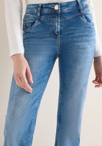 Jean en denim bleu avec poches avant et passants pour ceinture, porté avec un haut blanc à manches longues, mains détendues le long du corps.