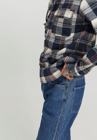 Camicia flanella a quadri blu navy e bianca con dettagli marroni, abbinata a jeans in denim azzurro chiaro con tasca posteriore. Look casual a strati.