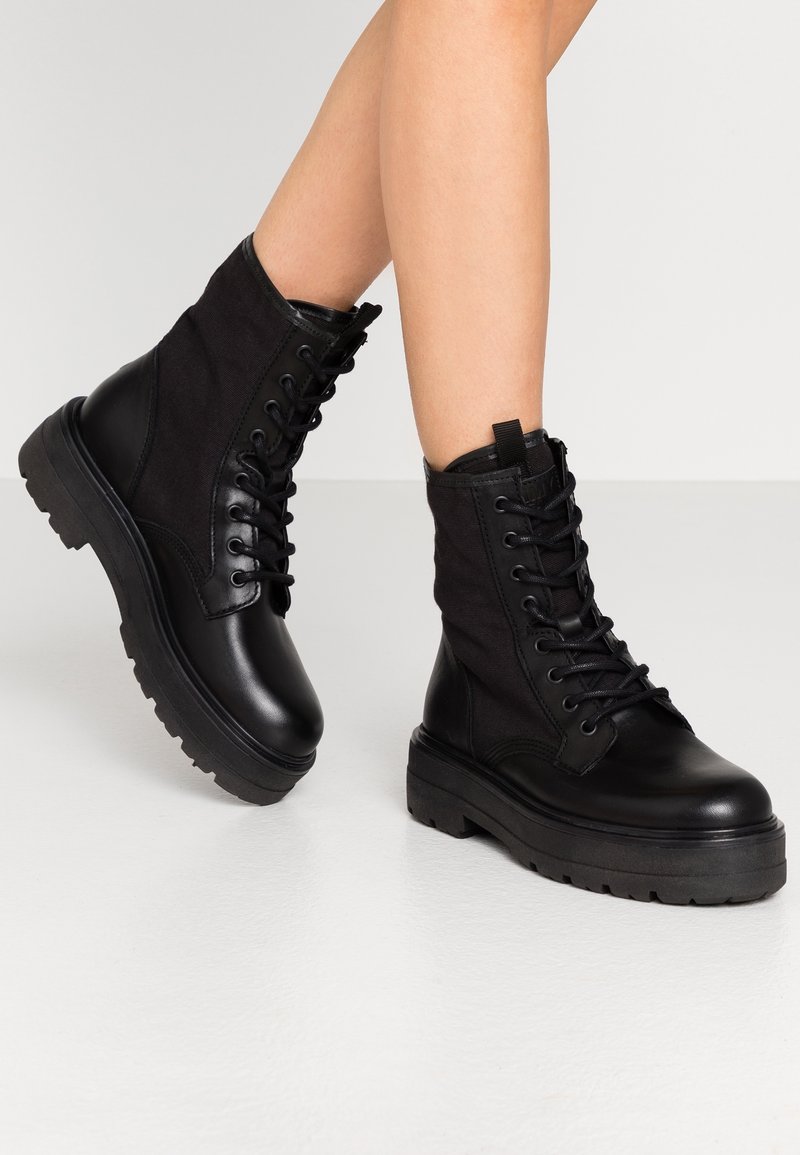 Schwarze Kampfstiefel aus einer Mischung von glattem Leder und strukturiertem Stoff, mit einer dicken Sohle und Schnürdesign.