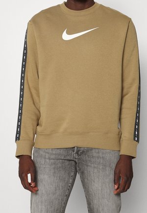 Mężczyzna w beżowej bluzie Nike z białym logo Swoosh na piersi i czarną taśmą z logo na rękawach, w połączeniu z szarymi dżinsami.
