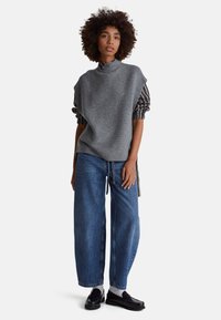 Žena s kudrnatými vlasy, oblečená v šedém svetru bez rukávů přes pruhovanou košili, volné modré džíny, bílé ponožky a černé mokasíny, stojí.