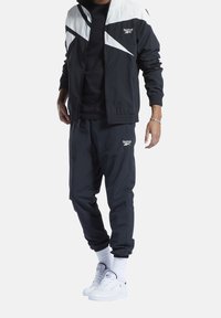 Man som bär en svart och vit Reebok träningsdräkt med logotyp, vita sockor av crew-modell och vita Reebok sneakers, står mot en enkel bakgrund.